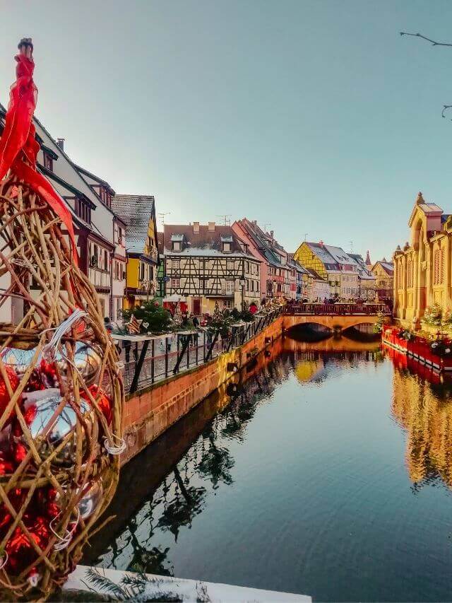 A river in Colmar, one of the best places to visit in winter in France, with houses covered in Christmas decorations and lights under clear blue skies.