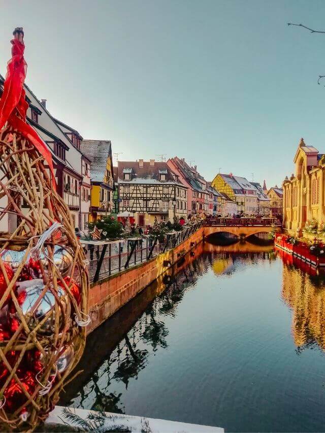 Un río en Colmar, uno de los mejores lugares para visitar en invierno en Francia, con las casas cubiertas de adornos y luces de Navidad bajo un cielo azul despejado.