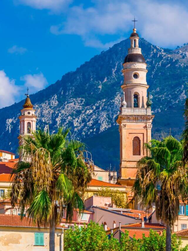 The towers of Menton church emerging from the roof of the surrounding houses with a view of snowy mountains,   making it one of the best places to visit in France in winter