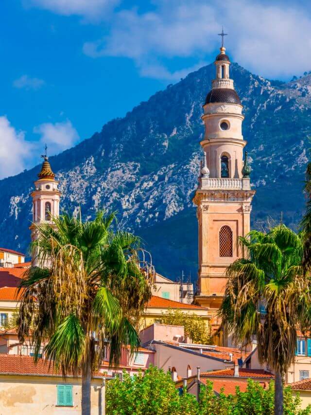 Las torres de la iglesia de Menton emergen del tejado de las casas circundantes con vistas a las montañas nevadas, lo que la convierte en uno de los mejores lugares para visitar en Francia en invierno (y también en verano).