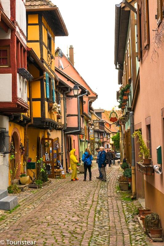 Una calle de Eguisheim con algunas personas paseando y hablando rodeadas de casas con plantas en macetas y poca decoración navideña, lo que la convierte en un destino mágico y en uno de los mejores lugares del invierno en Francia.