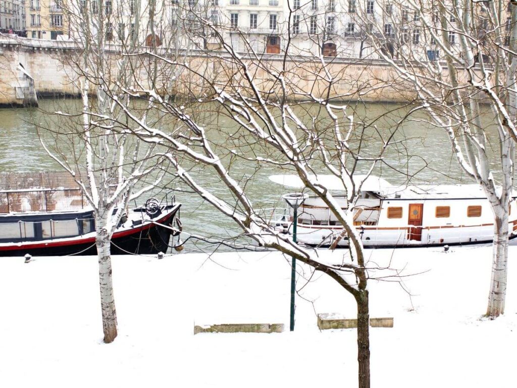 Boats docked on the side of the Seine River nearby walking path covered in snow in winter with a couple of trees. 
